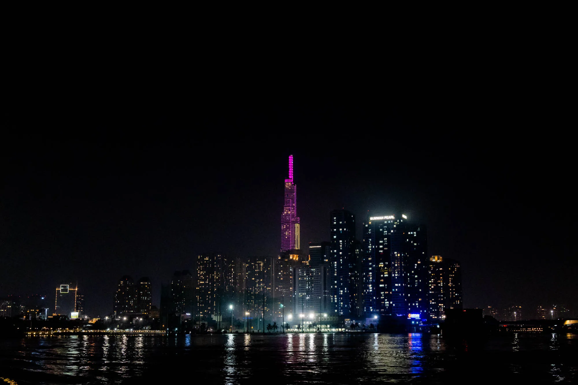 写真はベトナムのホーチミンで撮影された夜景です。漆黒の空を背景に、川面に反射する光が印象的です。特に目を引くのは、鮮やかなピンク色にライトアップされた高層ビルで、街のランドマークとなっているようです。その周りにも、多数のビルが光を灯しており、近代的な都市の風景を描き出しています。手前の川面は、光を反射してキラキラと輝き、夜景に奥行きを与えています。全体的に、都会的で洗練された雰囲気が感じられます。