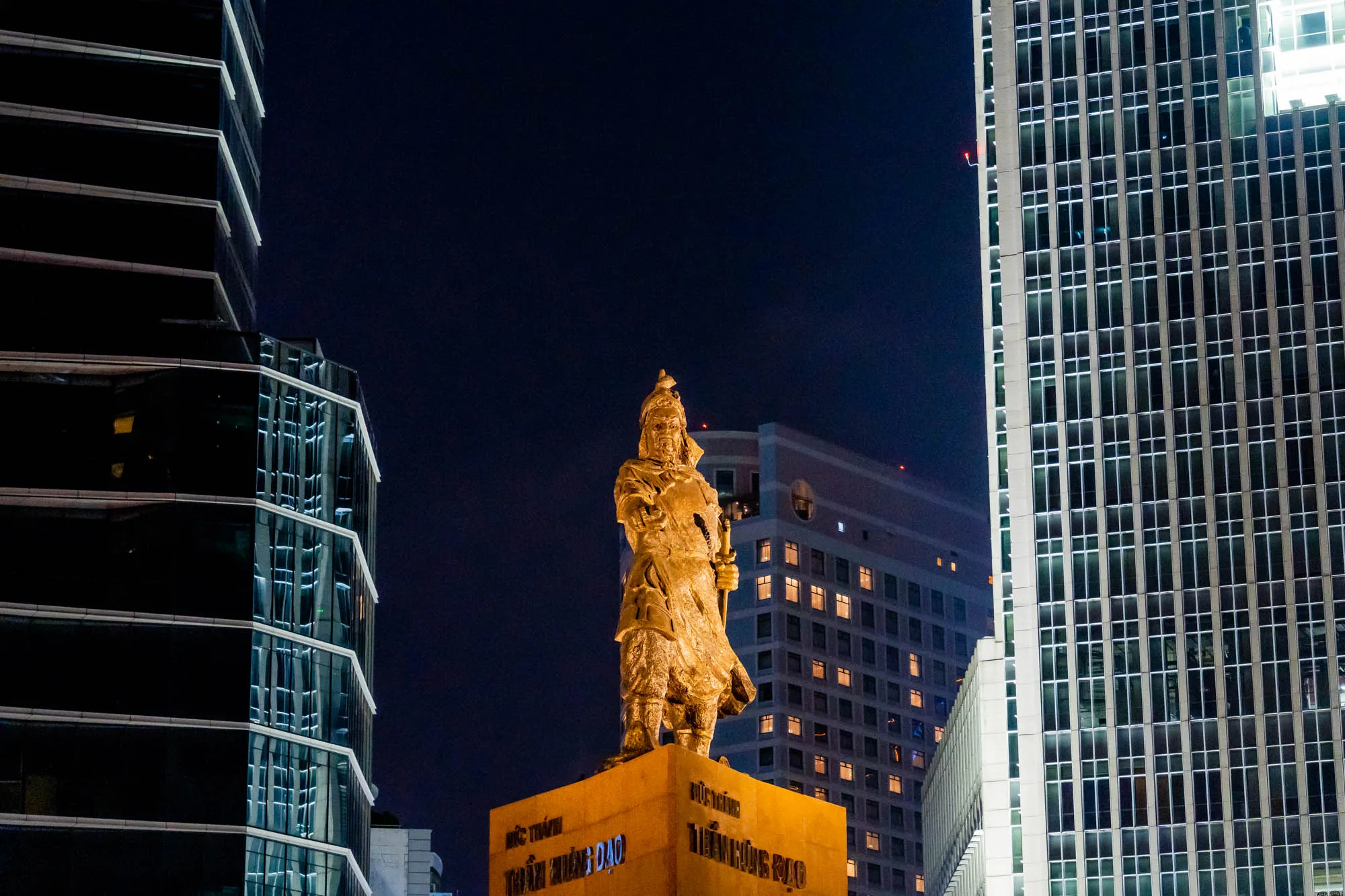 ホーチミンの夜に撮影された写真です。中央には黄金色にライトアップされた像が立っており、その背後には高層ビルがそびえ立っています。像は鎧を着た人物で、剣を持っているように見えます。像の土台には文字が刻まれています。左右には現代的なデザインのビルが写っており、窓から漏れる光が都会の雰囲気を醸し出しています。全体的に、伝統と現代が混ざり合った、ホーチミンの独特な風景を捉えた写真です。夜空は濃い青色で、ライトアップされた像とビルの光が際立っています。