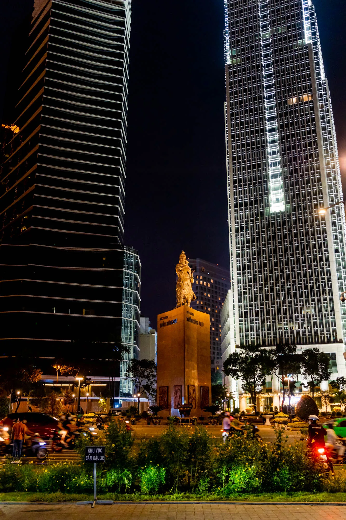 写真はベトナムのホーチミン市で撮影された夜景です。画面中央には、黄金色の像が立っており、それは大きな台座の上に設置されています。台座には文字が刻まれているようです。背景には、モダンな高層ビルが二棟そびえ立っています。左側のビルは黒く、横に走る白いラインが特徴的です。右側のビルは白く、縦横に走るラインが夜空に光を放っています。道路には、無数のバイクや車が走り、テールライトがぼやけて写っており、活気ある都市の様子を伝えています。手前には植え込みがあり、黄色い花が咲いています。全体的に、夜の街の賑やかさと、歴史的な像が組み合わさった印象的な光景です。