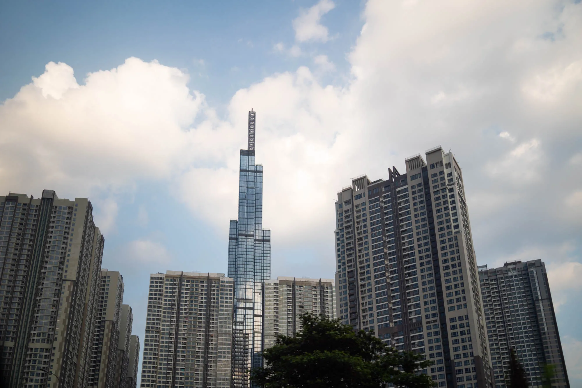 写真はベトナムのホーチミン市で撮影されたもので、複数の高層ビルが空に向かってそびえ立っている様子が捉えられています。中央には特に高い超高層ビルがあり、ガラス張りの外観が印象的です。その頂上には鉄骨構造のようなものが突き出ています。他の建物も高層マンションのようで、多くの窓が整然と並んでいます。空は晴れており、白い雲がいくつか浮かんでいます。手前には木々の緑が見え、都市景観に自然の要素を加えています。全体的に、ホーチミンの急速な都市開発と高層建築の進化を象徴するような風景です。