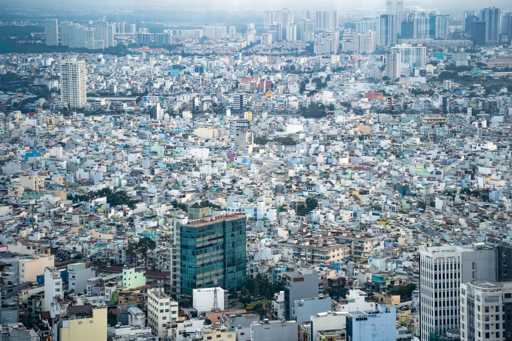 この写真は、ベトナムのホーチミン市を上空から撮影したものです。画面全体に、建物が密集して立ち並び、都市の広がりを感じさせます。手前には比較的低層の建物が密集し、その向こうには高層ビル群が広がっています。手前の建物は、パステルカラーの色使いが特徴的で、白、水色、薄緑色など、様々な色が混ざり合っています。高層ビル群は、白やグレーを基調としたモダンなデザインのものが多く、都市景観にコントラストを与えています。全体的に霞がかかったような雰囲気で、遠景はややぼやけて見えます。写真からは、ホーチミン市の活気と発展が感じられます。