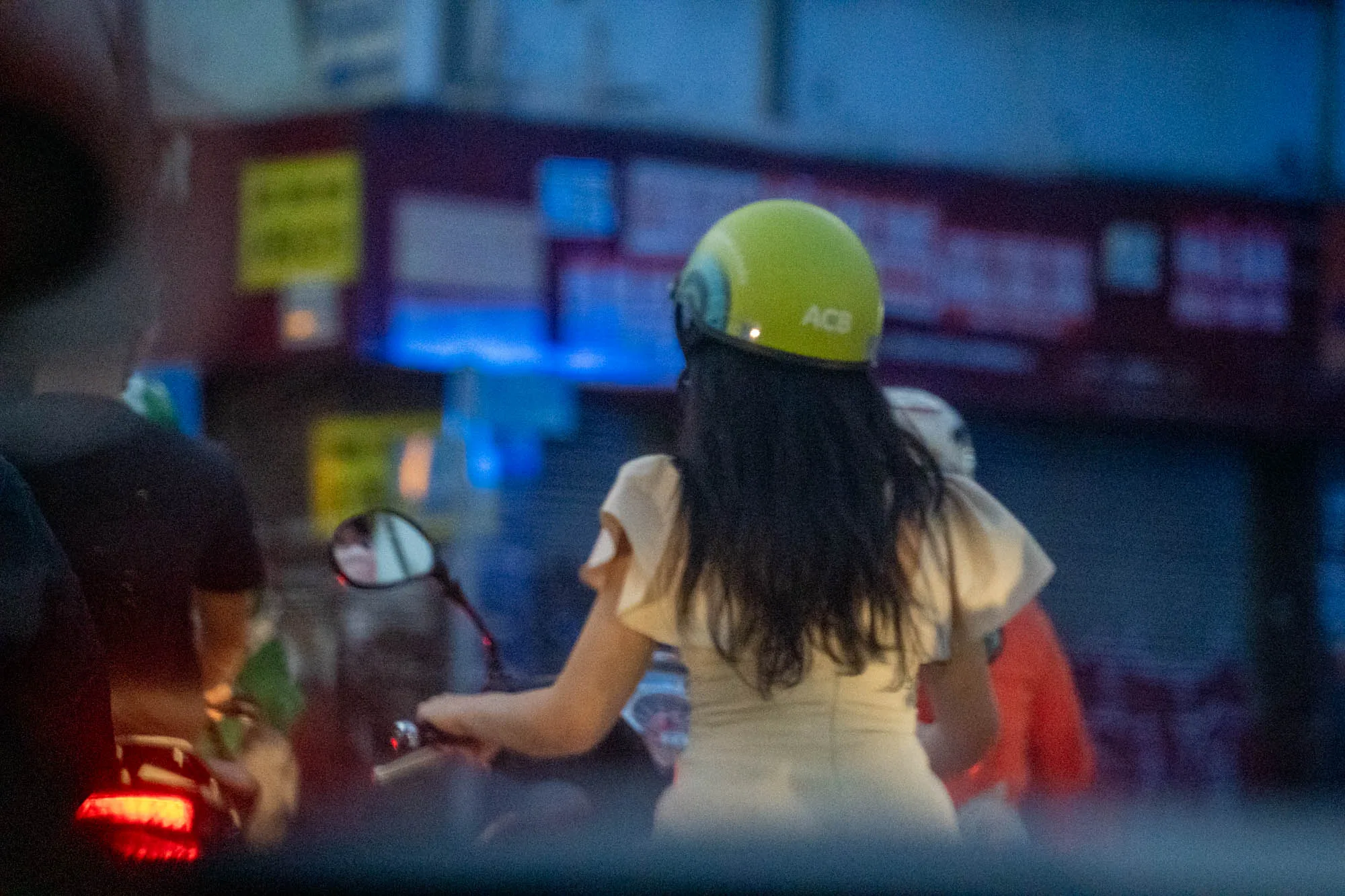 ホーチミンで撮影されたと思われる写真です。暗めの写真ですが、女性がバイクに乗っている様子が写っています。女性はクリーム色のワンピースのようなものを着ており、頭には蛍光色のヘルメットを被っています。ヘルメットには「ACB」という文字が見えます。女性の長い黒髪が背中に垂れています。  背景にはぼやけた看板やネオンサインのような光が見え、都市の夜の雰囲気を感じさせます。他にもバイクに乗った人が写っており、バイクの赤いテールランプが光っています。全体的に少しノイズが多く、暗い印象です。