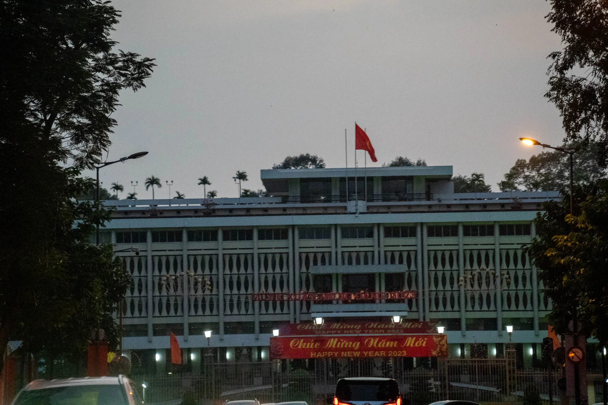 ベトナムのホーチミン市で撮影された写真です。画面中央に大きな建物が写っており、その屋上にはベトナム国旗が掲げられています。建物は複数の階があり、特徴的な縦長の窓が並んでいます。建物の正面には、「CHÚC MỪNG NĂM MỚI HAPPY NEW YEAR 2023」と書かれた赤い横断幕が掲げられています。これはベトナム語で「新年おめでとう」という意味です。建物の手前には道路があり、自動車が数台写っています。道路の両側には街路樹が植えられており、夕暮れ時の薄暗い空と相まって、落ち着いた雰囲気を醸し出しています。全体的に、ベトナムの新年を祝う静かで穏やかな情景が捉えられています。