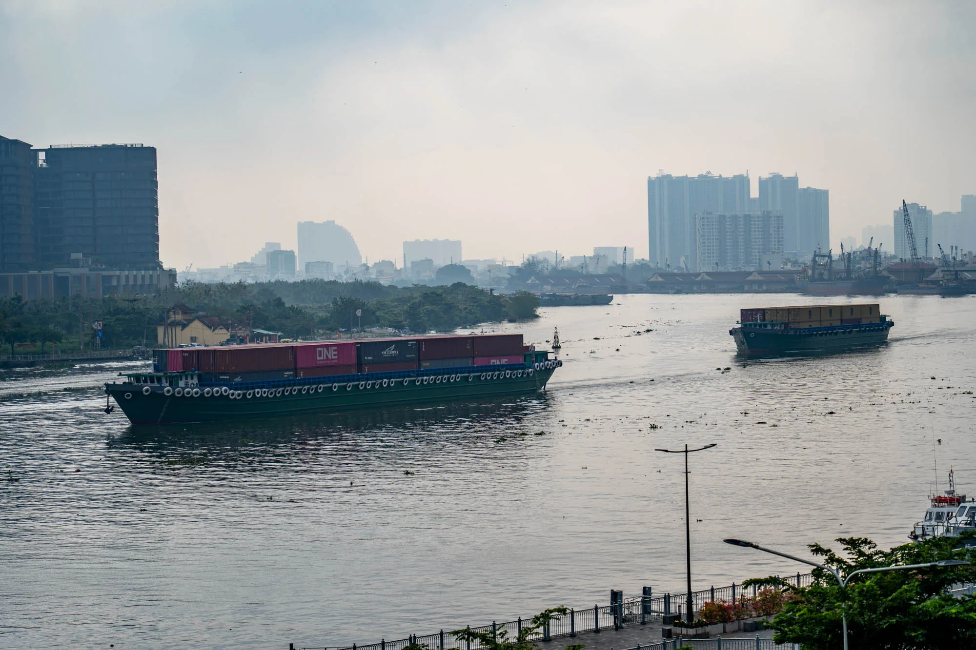 写真はベトナムのホーチミン市で撮影されたものです。手前には川が流れ、その上を2隻の平底船（バージ船）が航行しています。1隻は多くのコンテナを積んでおり、そのうちのいくつかには「ONE」と書かれたロゴが見えます。もう1隻は小型で、2層にコンテナが積まれています。背景には高層ビル群が立ち並んでおり、霧がかかっているのか、やや霞んで見えます。川岸には緑地が整備されており、街灯や遊歩道らしきものが見えます。全体的に穏やかな雰囲気で、都市の物流を支える水上輸送の様子が捉えられています。