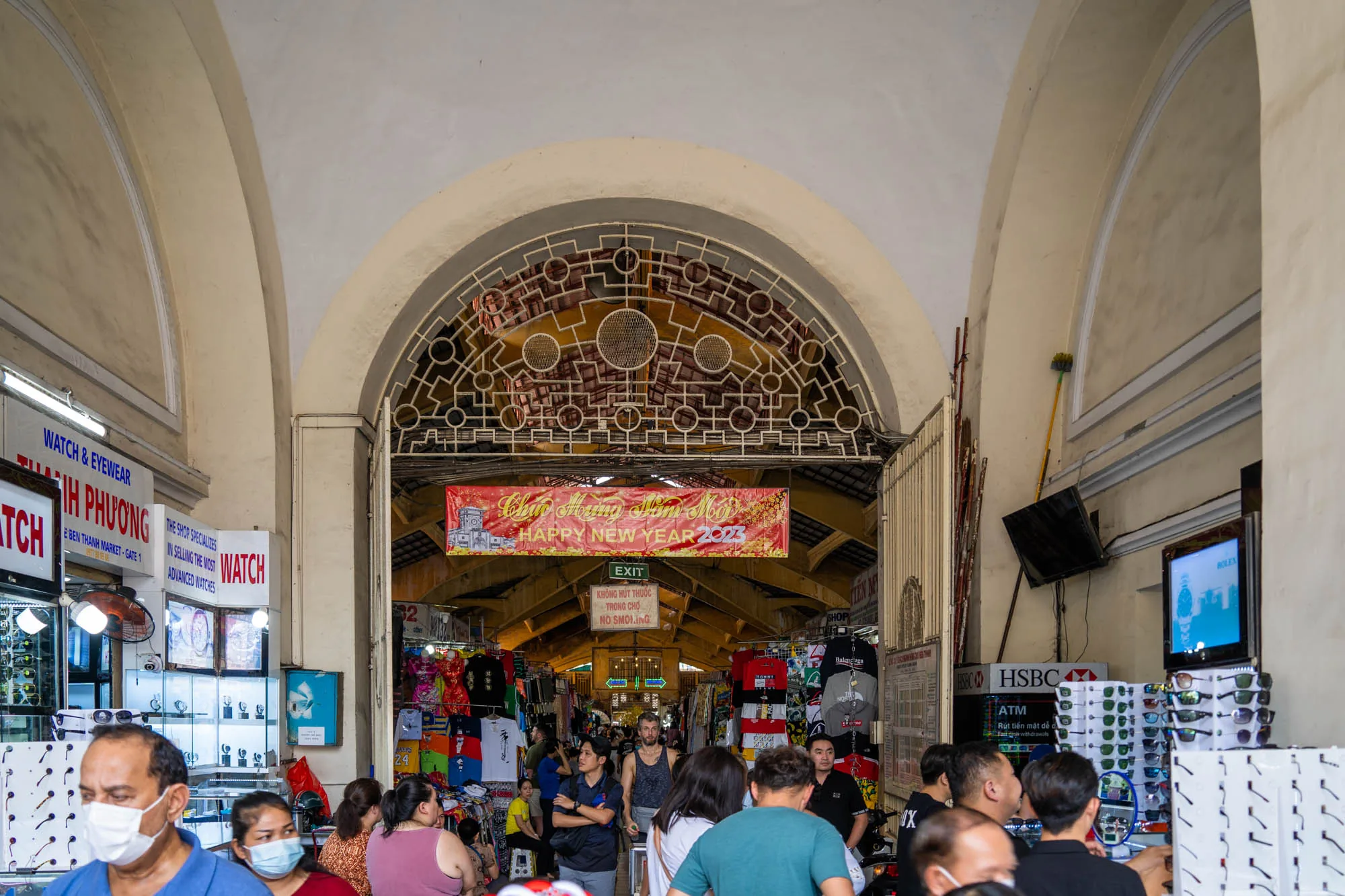 写真はベトナムのホーチミンにあるベンタイン市場の内部を捉えたものです。アーチ状の入り口から市場の様子が見え、多くの人々が行き交い、活気にあふれています。入り口の上には「Happy New Year 2023」と書かれた横断幕が掲げられており、撮影時期が2023年の新年であることを示唆しています。  市場内は様々な商品を扱う店がひしめき合っており、衣服や土産物などが並んでいます。入り口付近には時計や眼鏡を扱う店があり、商品がディスプレイされています。ATMの看板やテレビも見え、市場の機能が充実していることが伺えます。  全体的に、市場の喧騒と熱気が伝わってくる写真です。入り口の装飾や看板など、細部にも注意が払われており、市場の歴史や文化を感じさせます。人々の服装や表情から、日常生活の様子が垣間見え、活気ある市場の雰囲気が表現されています。