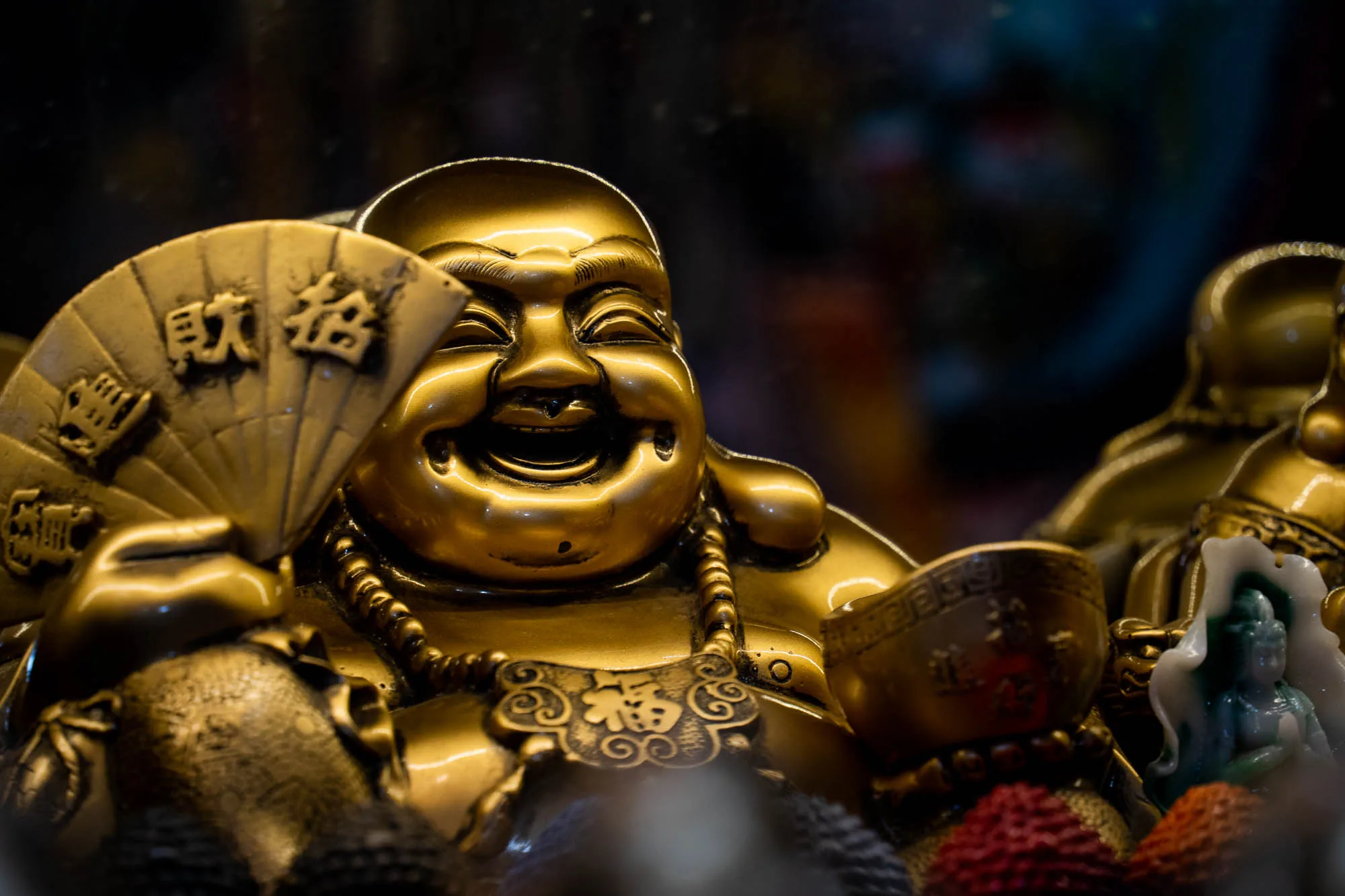 ホーチミンで撮影されたと思われる、金色の布袋像の写真です。布袋様は満面の笑みを浮かべ、扇子と宝珠を持っているように見えます。扇子には漢字が書かれており、財や福を招く意味合いが込められているようです。首飾りや胸元にも装飾が施されており、富を象徴するようなデザインです。背景は暗くぼやけており、布袋像の存在感が際立っています。他の像や装飾品も少し見えており、仏具店の一角で撮影されたのかもしれません。全体的に金色の光沢が美しく、幸福感に満ちた雰囲気の写真です。