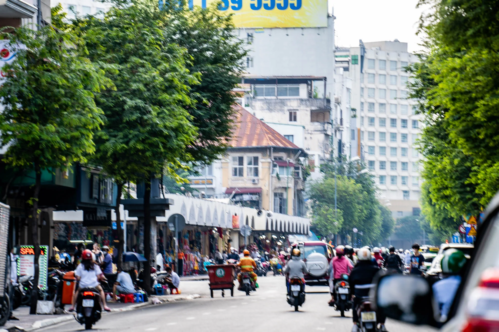 ベトナムのホーチミンの街並みを捉えた写真です。通りには多くのバイクが走り、活気にあふれています。歩道には露店や人々がいて賑やかです。背景には様々な建物が立ち並び、新旧の建物が混在している様子が伺えます。緑豊かな木々が街路樹として植えられており、都会の中にも自然を感じさせます。全体的に活気があり、エネルギッシュな雰囲気の写真です。