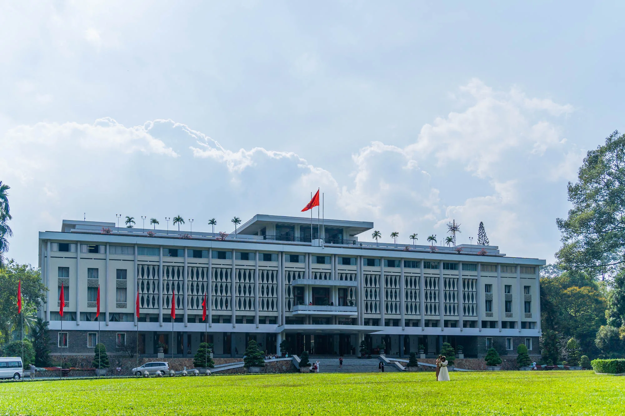 写真は、ベトナムのホーチミン市にある統一会堂（旧南ベトナム大統領官邸）を正面から捉えたものです。青空に白い雲が浮かび、建物の上にベトナム国旗が掲げられています。建物の正面には広い緑の芝生が広がり、手入れの行き届いた庭園が整備されています。数人の観光客らしき人々が建物の階段を上ったり、庭を散策している様子が見られます。奥には車が数台駐車されているのが見えます。全体的に穏やかで平和な雰囲気が漂っています。