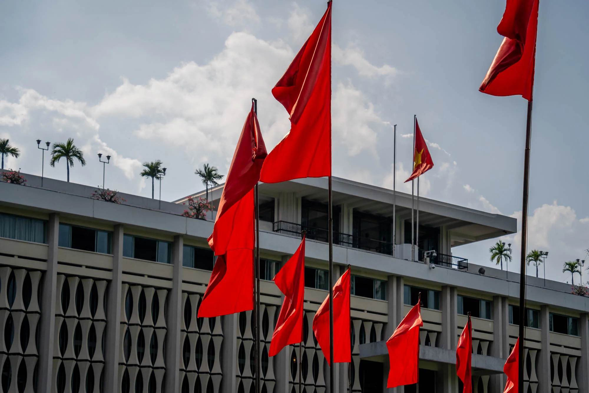 写真はベトナムのホーチミン市で撮影されたものです。前景には、鮮やかな赤色の旗が複数本、風になびいている様子が写っています。背景には、白い近代的な建物がそびえ立っており、その屋上にはヤシの木や照明が配置されています。建物の壁面は特徴的なデザインで、縦長の楕円形が連続して並んでいます。空は薄い水色で、雲がわずかに見られます。全体の雰囲気は、歴史的な場所でありながら、現代的な要素も感じられる、象徴的な風景です。