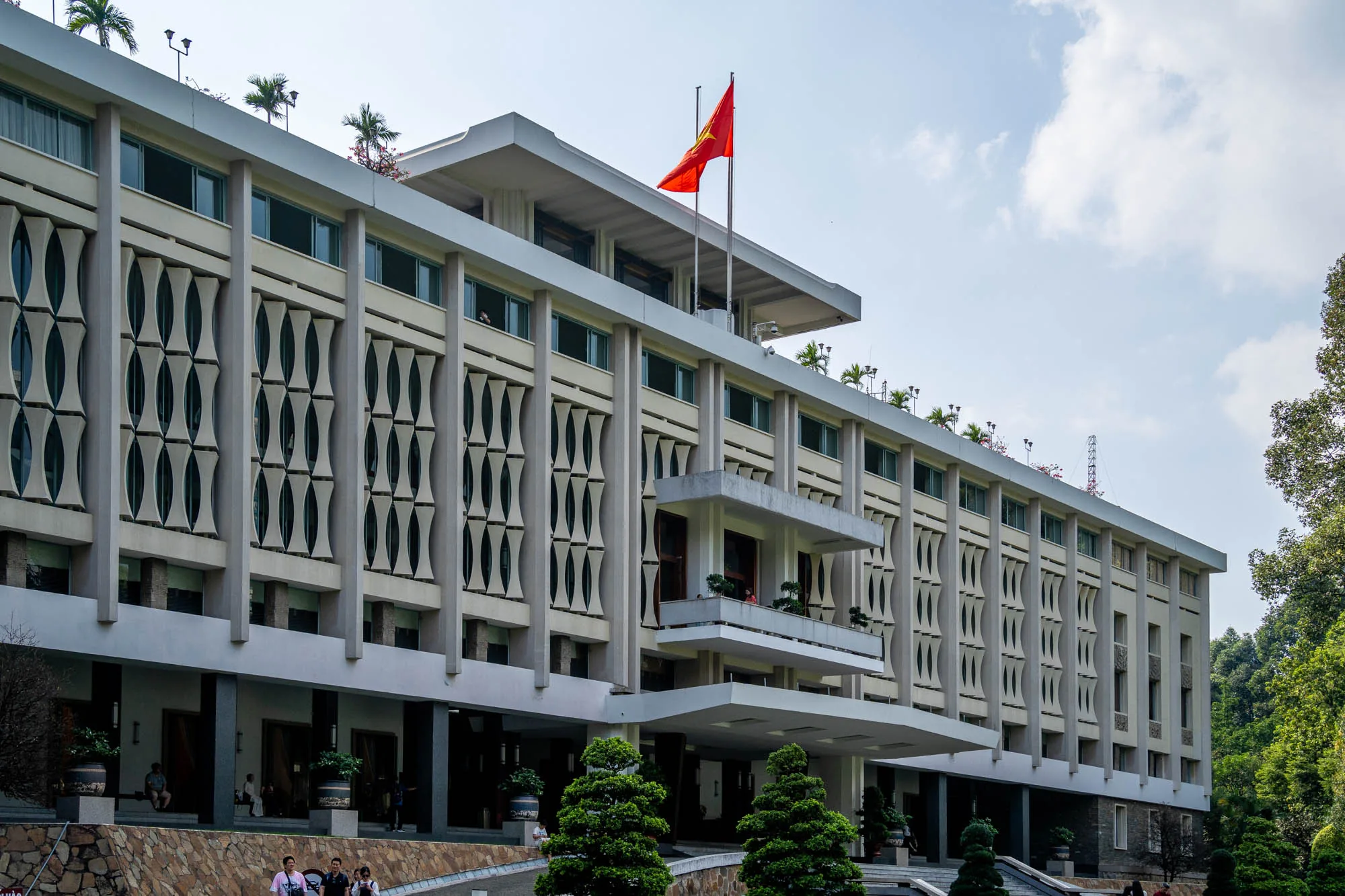 写真には、ベトナムのホーチミン市にあると思われる、大きな近代的な建物が写っています。建物の屋上にはベトナムの国旗が掲げられています。建物は白っぽい色で、独特の縦長の装飾が施された窓が並んでいます。建物は木々や緑に囲まれており、敷地内には人が数人いるのが見えます。空は一部曇っています。全体的に、威厳があり、歴史的な建造物である印象を与えます。