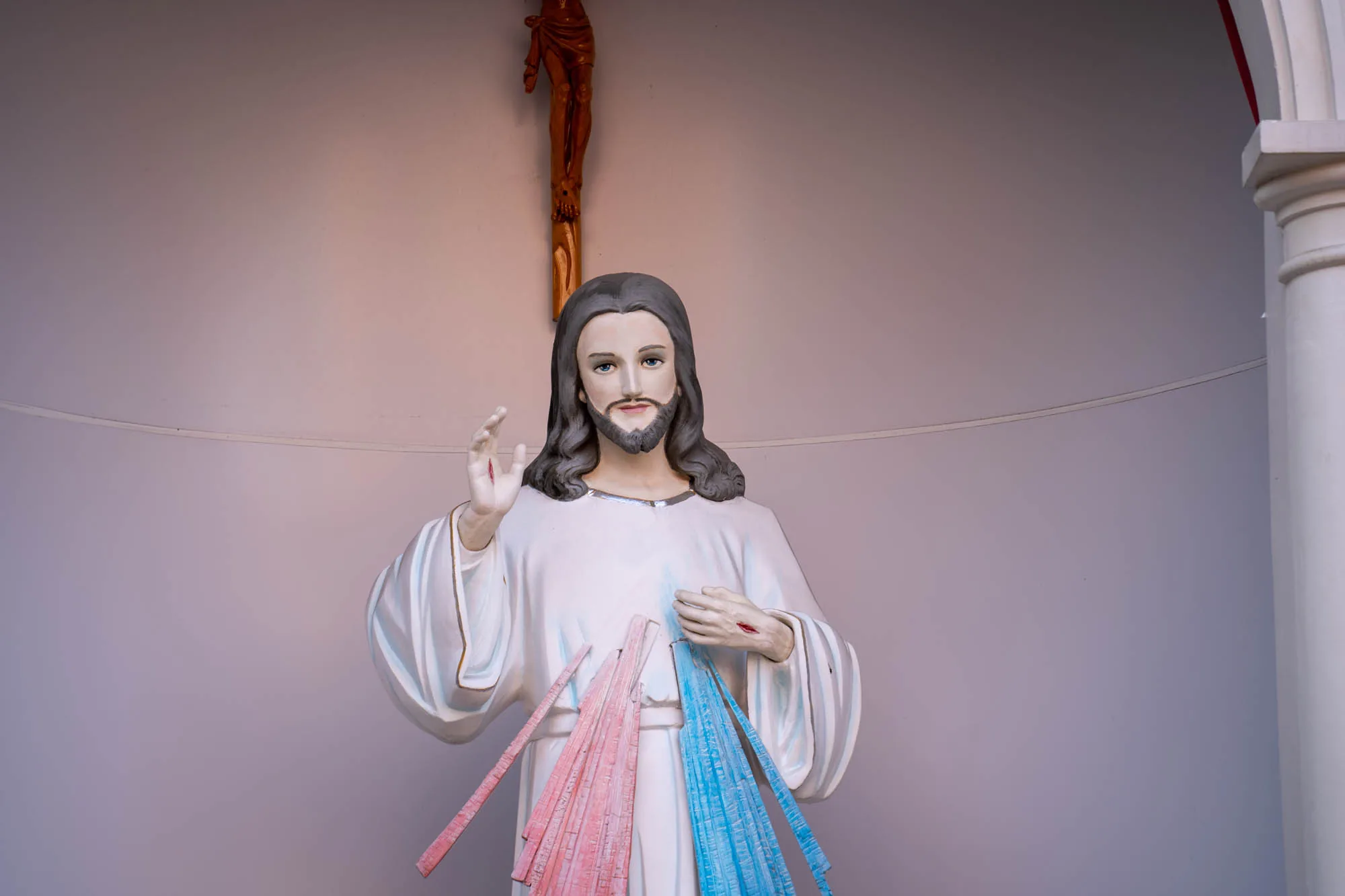 写真は、ベトナムのホーチミンで撮影されたイエス・キリスト像を捉えています。像は、白いローブを身に着け、傷のある手を広げて祝福しているように見えます。胸からは赤い光と青い光のようなものが放射されています。背景には木製の十字架に磔にされたイエス像が見えます。全体的に、神聖で敬虔な雰囲気を醸し出しています。像は壁の前に置かれ、右側には白い柱の一部が見えます。