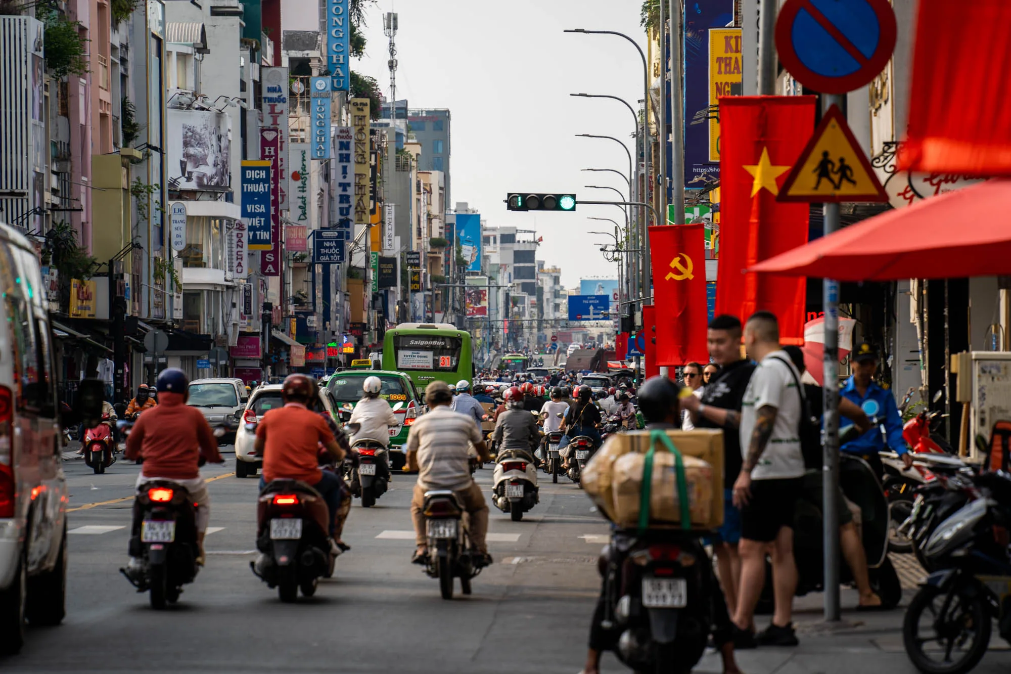 この写真はベトナムのホーチミンで撮影されたもので、交通量の多い街の様子を捉えています。画面中央には、多数のバイクが道路を埋め尽くし、前へ進もうとしているのが見えます。ほとんどの人がヘルメットを着用しており、様々な色の服を着ています。バイクの中には荷物を積んでいるものもあります。  道路の両側には、色とりどりの建物が立ち並び、多くは看板や広告で覆われています。ベトナム語の看板が多く、様々な商品やサービスを宣伝しているようです。電柱や電線が空を覆い、都市の雑然とした雰囲気を強調しています。  写真右側には、ベトナムの国旗が並び、赤い傘のある屋台や人が立っています。赤はベトナムを象徴する色のようで、街の活気とエネルギーを感じさせます。  全体的に、この写真はホーチミンの喧騒とした日常と、その独特な都市景観を伝えています。交通渋滞や建物の密集具合から、活気あふれるアジアの大都市であることがわかります。