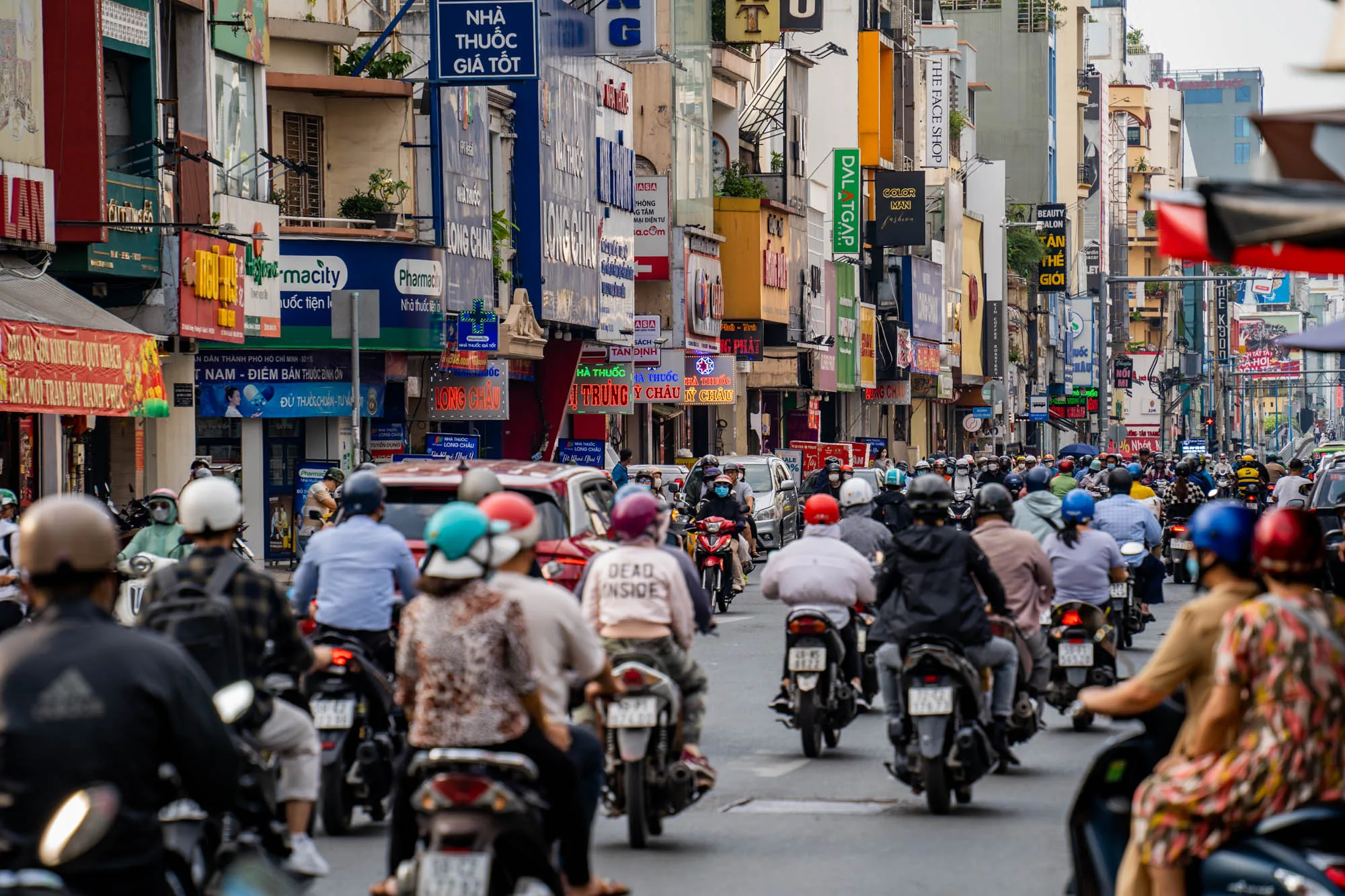 写真はベトナムのホーチミン市で撮影されたもので、バイクの群れが道路を埋め尽くしている様子が写っています。手前から奥へと続く道路には、無数のバイクがひしめき合っており、そのほとんどの運転手がヘルメットを着用しています。バイクの間に時折自動車も確認できます。背景には、高さの異なる建物が立ち並び、カラフルな看板や広告が所狭しと掲げられています。これらの看板はベトナム語で書かれており、さまざまな商店や企業を宣伝しています。空はやや曇っており、埃っぽくも感じられ、都会の喧騒と活気が伝わってきます。全体的に、ホーチミンの交通量の多さと、独特の都市景観が捉えられた一枚と言えるでしょう。
