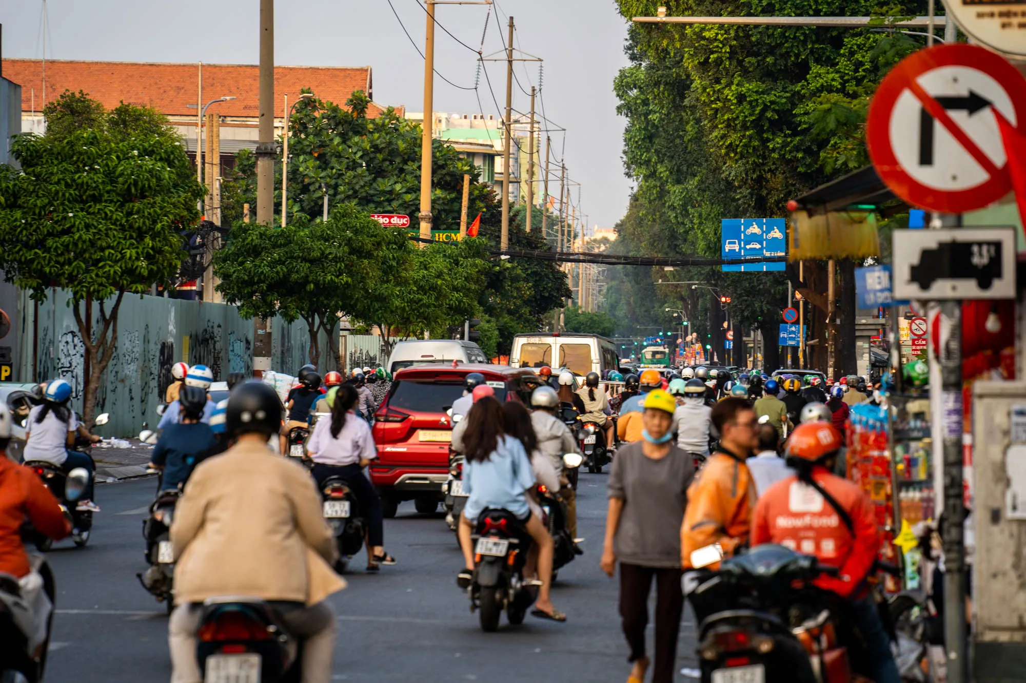 ベトナムのホーチミン市で撮影された写真です。写真には、道路を埋め尽くすように多くのバイクが写っています。それぞれのバイクには運転手や乗客が乗っており、様々な服装をしています。多くがヘルメットを着用しています。道路は混雑しており、交通量が多いことがうかがえます。道路の脇には建物や木々が立ち並び、電線が空中に張り巡らされています。右側には交通標識がいくつか見えます。背景には、建物や木々が連なり、ベトナムの都市風景が広がっています。全体的に活気があり、エネルギーに満ちた雰囲気の写真です。