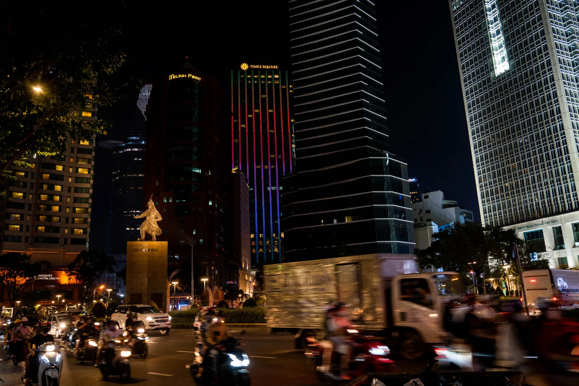 この写真はベトナムのホーチミン市で撮影された夜景です。画面中央には、カラフルにライトアップされた高層ビル群がそびえ立ち、「MELINH POINT」や「TIMES SQUARE」といった文字が見えます。これらの高層ビルは、夜の都市景観を華やかに彩っています。 写真の下部には、バイクや車が多数行き交う様子が写されており、活気のあるホーチミンの夜の交通状況を捉えています。手前にはバイクのヘッドライトが輝き、背景の建物群と相まって、都会の喧騒とエネルギーを感じさせます。 また、画面左側には像が建てられており、歴史的な要素も感じられます。写真全体を通して、ホーチミン市の現代的な発展と伝統的な魅力が共存している様子が伝わってきます。夜の暗さと建物の光のコントラストが印象的な一枚です。