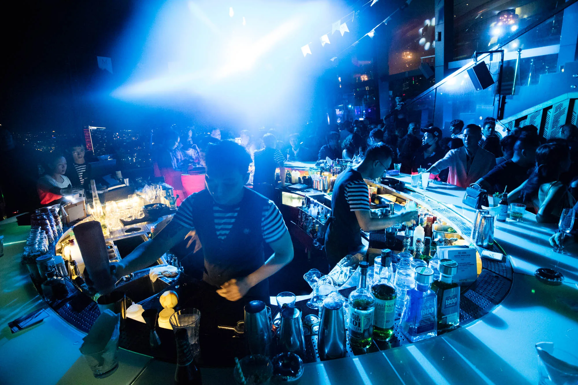 写真はベトナムのホーチミンで撮影されたと思われる、賑やかなナイトバーの様子を捉えています。画面全体が強い青色の光で照らされており、幻想的で活気のある雰囲気を醸し出しています。  手前には、バーカウンターがあり、ストライプのシャツを着たバーテンダーが2人、カクテルを作っている様子が見えます。カウンターには様々な種類のボトルやグラス、シェーカーなどが並べられ、プロフェッショナルな雰囲気が漂っています。  カウンターの向こう側には、多くの人々が集まり、お酒を飲んだり会話を楽しんだりしている様子が伺えます。暗い背景にはホーチミンの夜景が広がり、高層ビルなどの明かりがぼんやりと見えています。  写真全体を通して、ナイトライフの華やかさや活気が感じられ、ホーチミンの夜の魅力を伝えています。