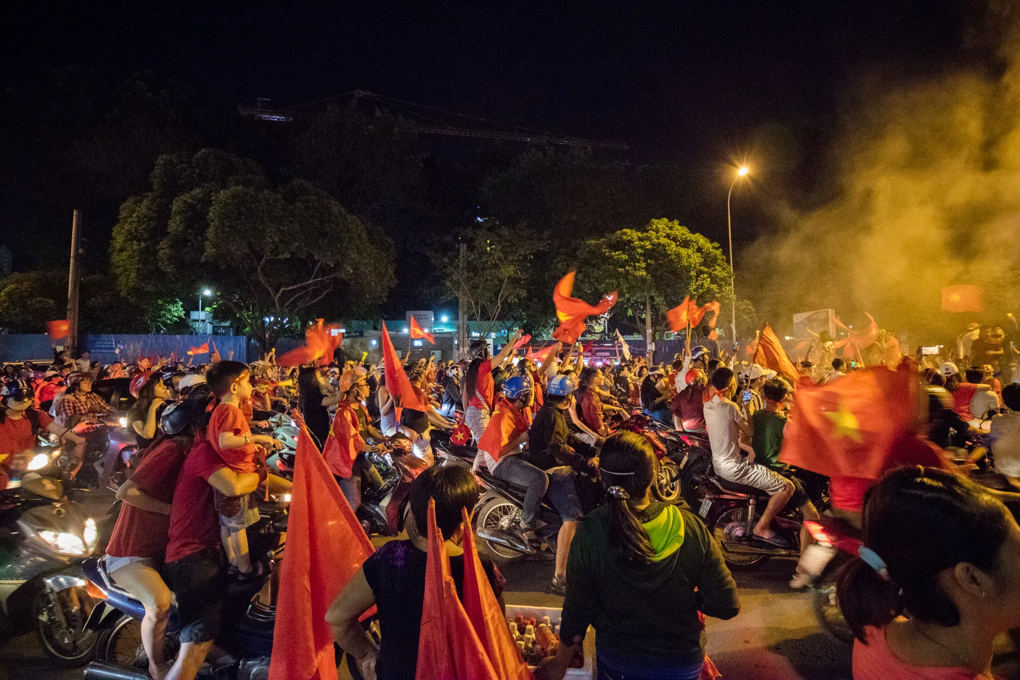 この写真はベトナムのホーチミン市で撮影された夜の風景です。大勢の人々がバイクに乗り、ベトナム国旗を掲げています。彼らは何らかのイベントや祝典を祝っているように見えます。夜であるため、周りは暗く、街灯やバイクのヘッドライトが光っています。背景には木々や建設中のクレーンが見えます。また、煙が立ち上っている場所もあり、お祝いの雰囲気をさらに盛り上げています。人々の表情は見えませんが、彼らの熱気が伝わってくるような、活気のある写真です。