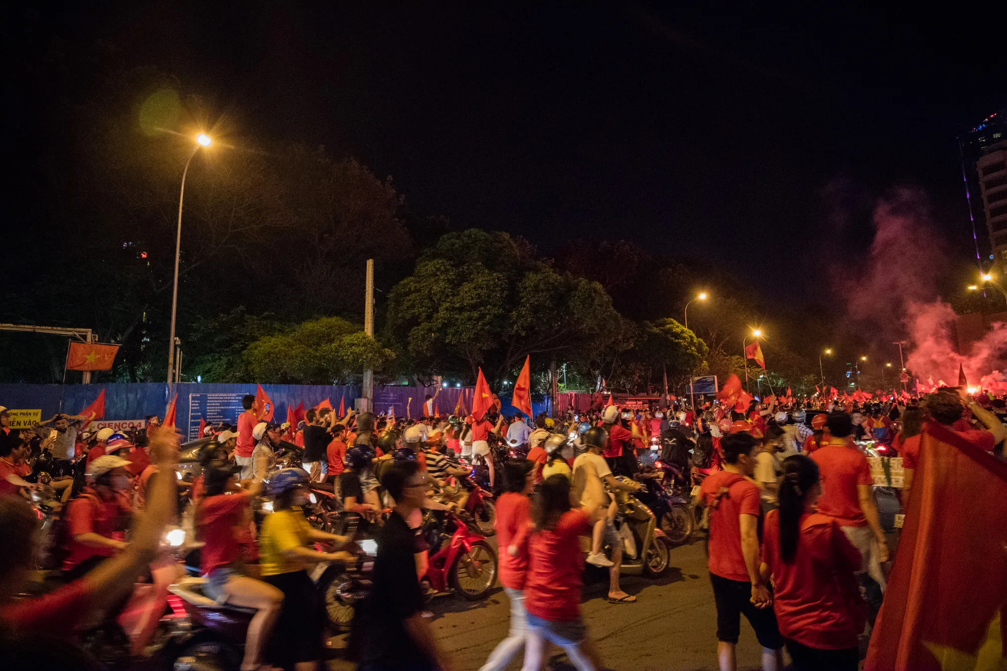 写真はベトナムのホーチミン市で撮影された夜の様子を捉えています。大勢の人々がベトナム国旗を掲げ、赤い服を着て、バイクに乗りながら歓声を上げているようです。集団は通りを埋め尽くし、熱気に満ち溢れています。背景には街灯や木々、建設現場のフェンス、高層ビルなどが写っています。一部には赤い煙が立ち上っており、祭りのような騒ぎであることがうかがえます。全体的にエネルギッシュで高揚感のある雰囲気が感じられます。