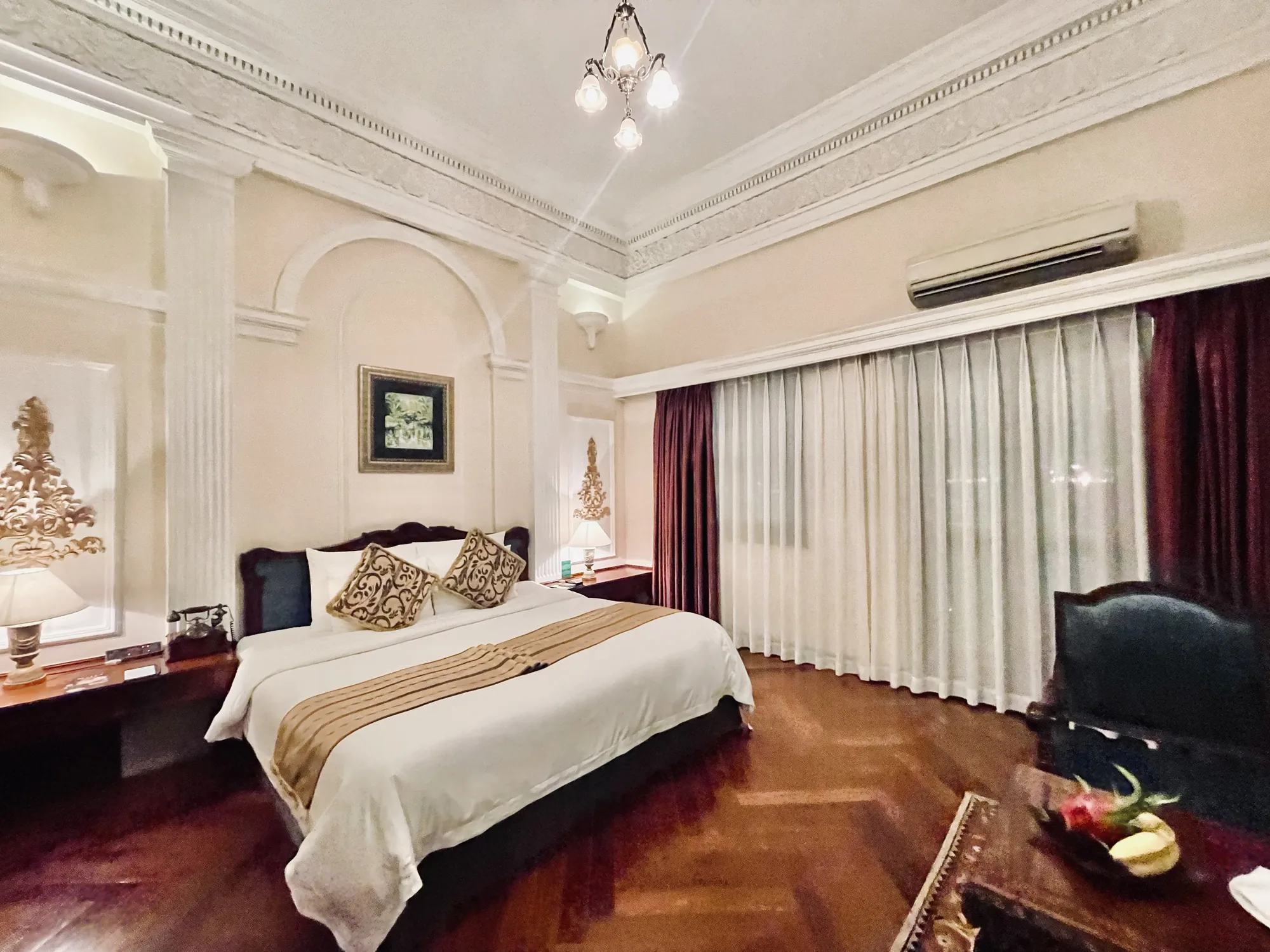 写真は、ベトナムのホーチミンにあるホテルの部屋の内部を写したものです。部屋はクラシカルな内装で、天井には豪華な装飾が施され、シャンデリアが吊り下げられています。壁はクリーム色で、アーチ状の装飾や柱が施されています。部屋の中央にはキングサイズのベッドが置かれ、白いリネンにゴールドのベッドランナーがかけられています。ベッドサイドには木製のナイトテーブルがあり、ランプやアンティーク調の電話が置かれています。窓には白いレースのカーテンと、濃い赤色のドレープカーテンが掛けられています。部屋の奥には、黒い革張りの椅子と、フルーツが盛られたテーブルがあります。床は濃い茶色の木製フローリングで、照明の反射で光沢があります。全体的に落ち着いた雰囲気で、高級感のある空間です。