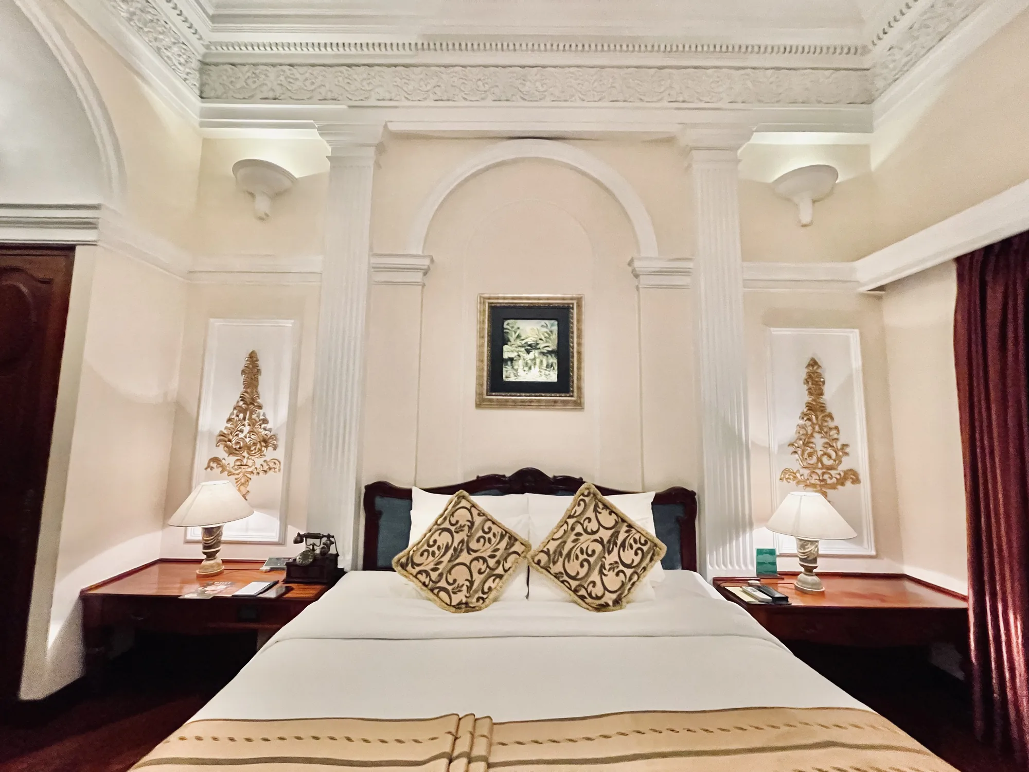 ホーチミン市で撮影されたホテルの客室の写真です。写真は、部屋の中心に置かれたベッドを正面から捉えています。ベッドは白いシーツと、茶色とベージュの模様が入ったクッションで飾られています。ベッドのヘッドボードは濃い青色で、落ち着いた印象を与えます。ベッドの両側には木製のナイトテーブルがあり、それぞれにランプが置かれています。左側のナイトテーブルには、古いスタイルの電話機が置かれています。壁はクリーム色で、アーチ状のデザインと白い柱が施されています。壁には絵画と装飾的な金色の彫刻が飾られています。部屋全体は、クラシックでエレガントな雰囲気です。