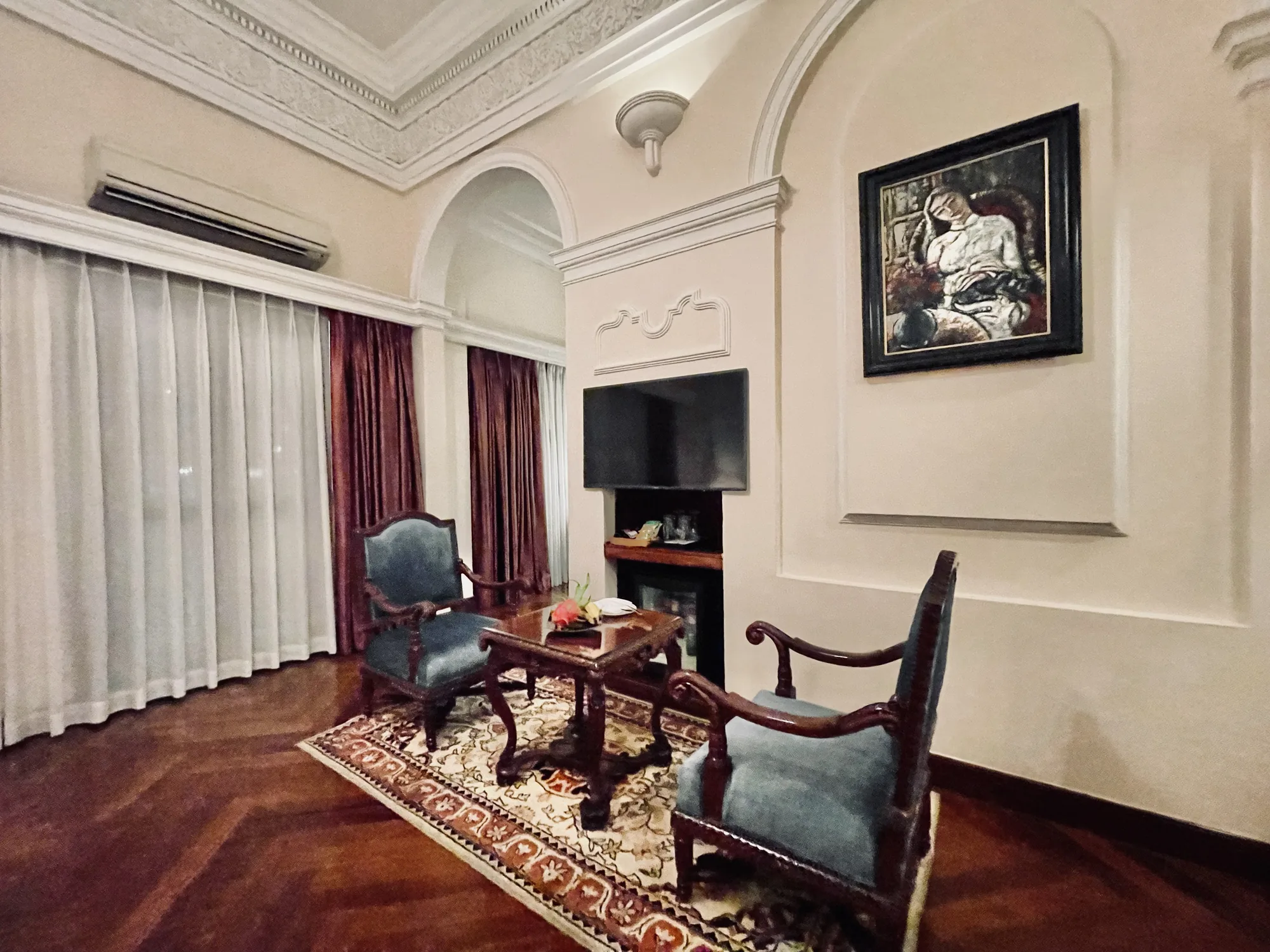 写真には、ベトナムのホーチミンにある豪華なホテルの客室の一部が写っています。室内はクリーム色の壁で、天井には凝った装飾が施されています。床は木製の寄木張りで、上質な雰囲気があります。  窓には白いシアーなカーテンと、光沢のある赤茶色の遮光カーテンが重ねてかけられています。部屋の中央には、花や果物が飾られた木製のコーヒーテーブルがあり、その周りを青いベルベットの肘掛け椅子が囲んでいます。  壁にはテレビが埋め込まれており、その上には装飾的な額縁が施されています。さらに、古典的な肖像画が飾られています。部屋全体が上品で落ち着いた雰囲気です。