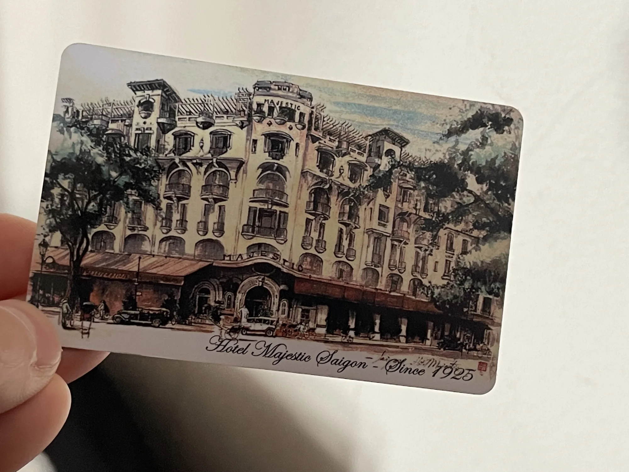 写真には、指でつまんだ絵葉書が写っています。絵葉書には、ベトナムのホーチミンにあるマジェスティックホテルの絵が描かれています。ホテルは歴史を感じさせる外観で、「Hotel Majestic Saigon - Since 1925」と書かれています。絵は水彩画のようなタッチで描かれており、レトロな雰囲気を醸し出しています。ホテル前には車や人が描かれ、活気のある様子が表現されています。
