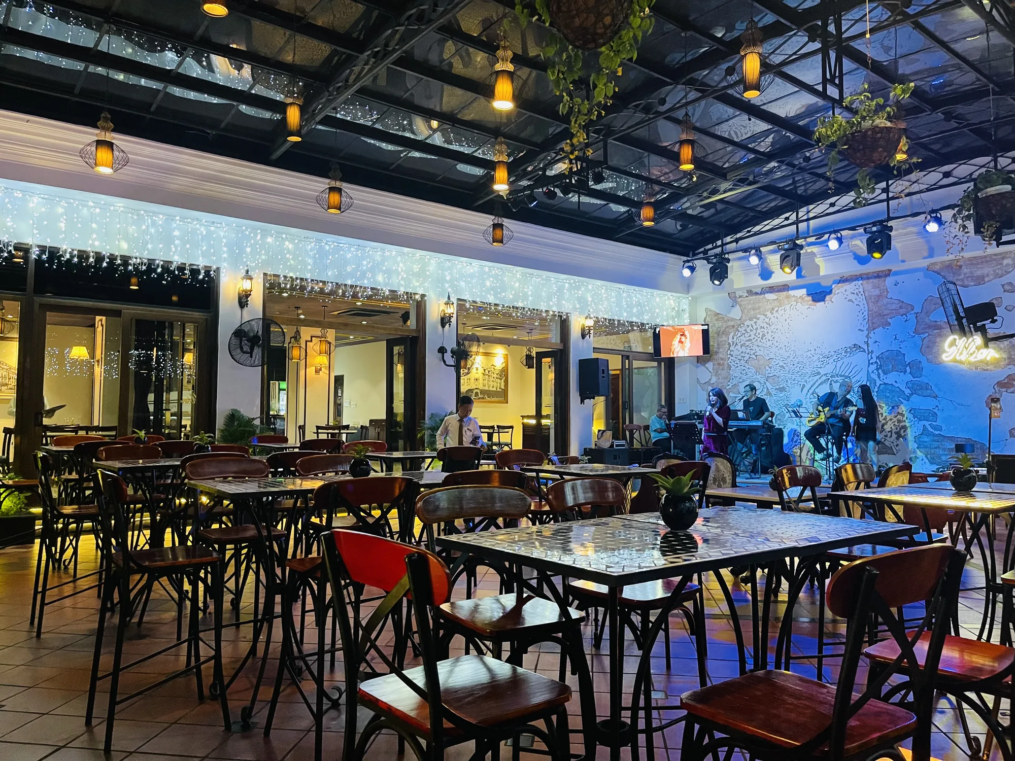 写真はベトナムのホーチミンで撮影されたライブレストランの様子を捉えています。店内はテーブルと椅子が並べられ、ステージではバンドが演奏しています。天井はガラス張りで、シャンデリアのような照明とガーランドライトが飾られ、雰囲気のある空間を作り出しています。ステージの背景には壁画が描かれ、ライトアップされています。店内には、お客さんとウェイターの姿が見えます。全体的に、落ち着いた雰囲気で、音楽を楽しみながら食事やお酒を楽しめる場所であることが伺えます。